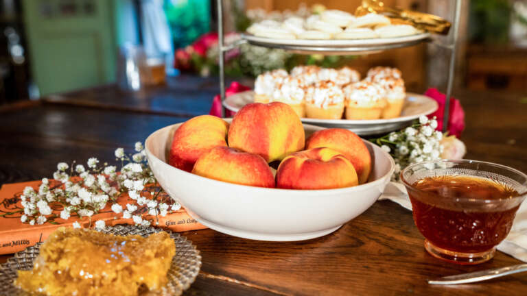 A tea party table set with a plate of honeycomb, a tiered tray stand laden with pastries, a bowl of peaches and baby's breath and antique books as part of the theme's decorations.