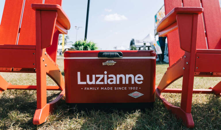 two red adirondack chairs with a red luzianne tea cooler in between them, on a sunny day at a festival